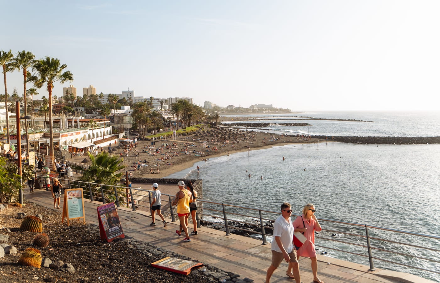 People walking the seafront promenade at Playa de las Américas past palms and low buildings